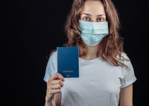 Reizen voor de covid-19 pandemie. Vrouw in een medisch masker houdt een paspoort op een zwarte muur.