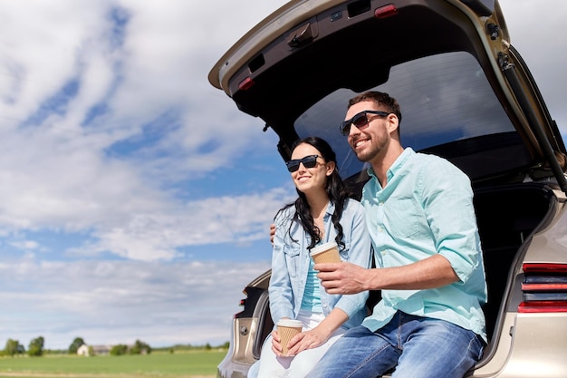 reizen, zomervakantie, road trip, vrije tijd en mensen concept - gelukkige paar koffie drinken uit wegwerpbekers zittend op kofferbak van hatchback auto buitenshuis