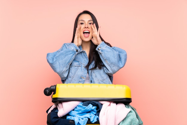 Reiziger Colombiaanse vrouw met een koffer vol kleren over geïsoleerde roze muur schreeuwen met wijd open mond