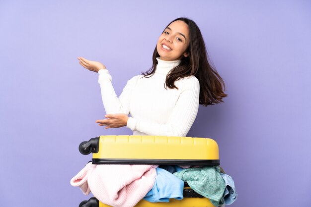 Reiziger jonge vrouw met een koffer vol kleren over paars uitgestrekte handen opzij om uit te nodigen om te komen