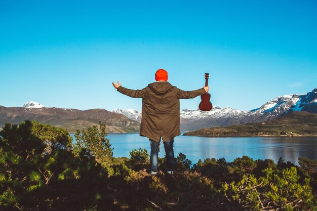 Reiziger man staat in het midden van een bos met een gitaar op de achtergrond van bergen en meer