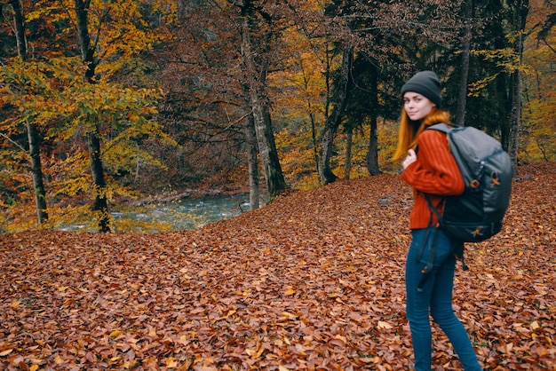 Reiziger met een rugzak in het herfstbos en hoed, trui, jeans, gevallen bladeren, bomen van het meer