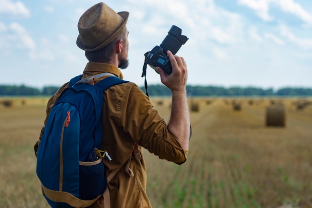 Reizigersfotograaf met een camera in zijn hand tegen de achtergrond van een veld en hooibergen.