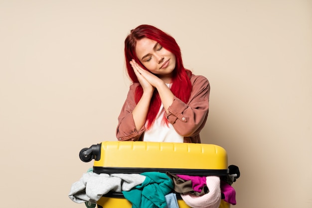 Reizigersmeisje met een kofferhoogtepunt van kleren op beige wordt geïsoleerd dat slaapgebaar dat in dorable uitdrukking maakt