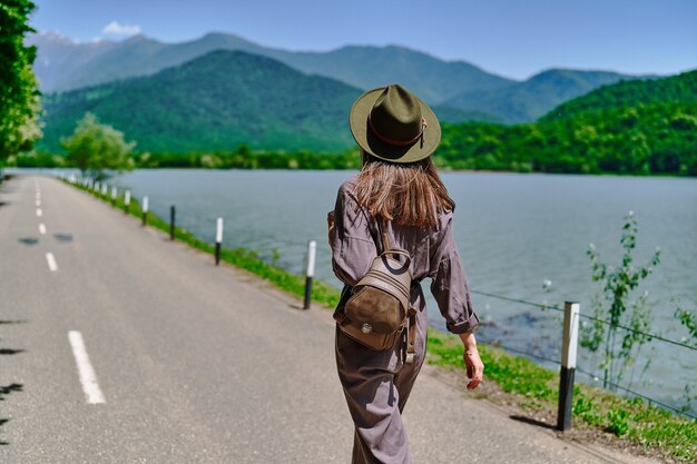 Reizigersmeisje met vilten hoed en rugzak die alleen op een weg loopt met uitzicht op het meer en de bergen. genieten van een prachtig vrijheidsmoment van het leven en een serene rustige vredige kalme sfeer in de natuur. achteraanzicht