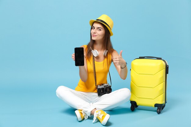 Reizigerstoeristenvrouw in gele vrijetijdskleding, hoed met koffer mobiele telefoon op blauw