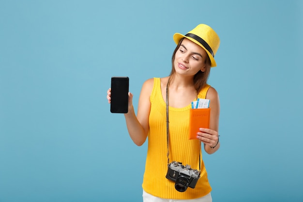 Reizigersvrouw in gele vrijetijdskleding en hoed met kaartjes, mobiele telefoon en camera op blauw