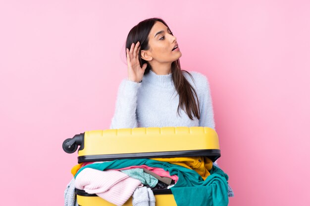 Reizigersvrouw met een kofferhoogtepunt van kleren over geïsoleerde roze muur die iets luisteren