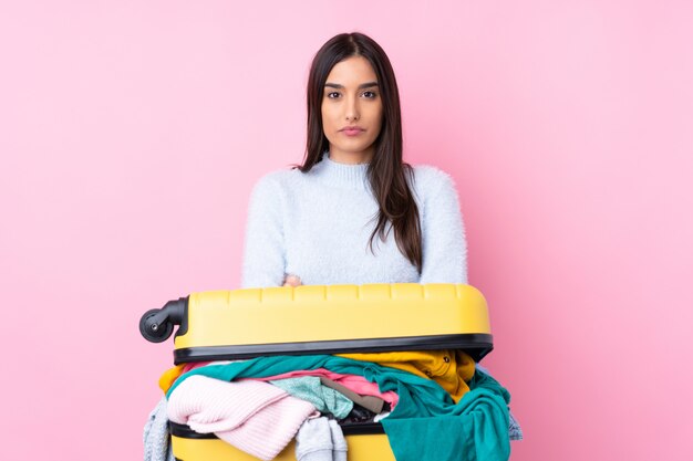 Reizigersvrouw met een kofferhoogtepunt van kleren over geïsoleerde roze muur die wapens gekruist houden