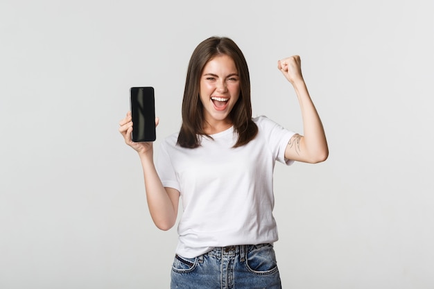 Rejoicing beautiful girl showing smartphone screen and dancing like champion.