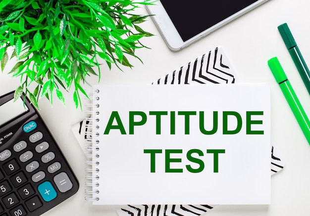 Rekenmachine, groene plant, telefoon, stift, notitieboekje met de tekst APTITUDE TEST op het bureaublad