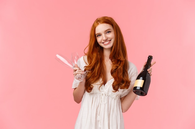 Relatie, feest en tederheid concept. Het mooie Kaukasische meisje met rood lang krullend haar in witte kleding, nodigt heeft samen drank uit, houdend champagne en twee glazen, het glimlachen