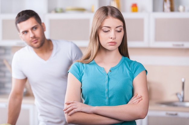 Relationship difficulties. Depressed young woman keeping arms crossed and looking away while man standing behind her