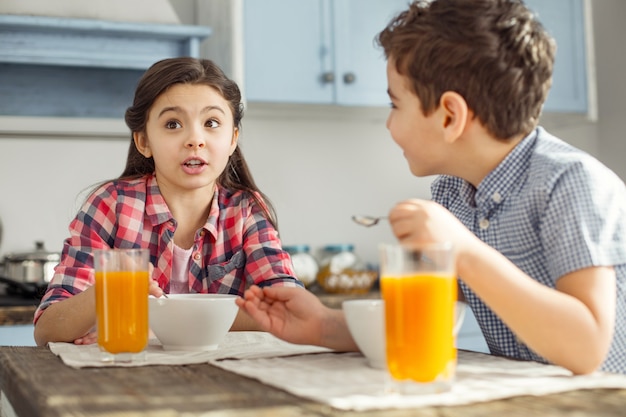 Relationship. Pretty alert little dark-haired girl talking with her brother while they having breakfast and drinking some juice