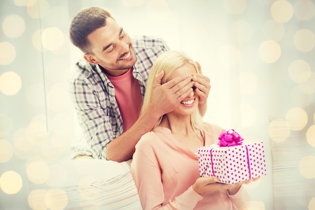 Photo relationships, love, people, birthday and valentines day concept - happy man covering woman eyes and giving gift box at home over holidays lights background