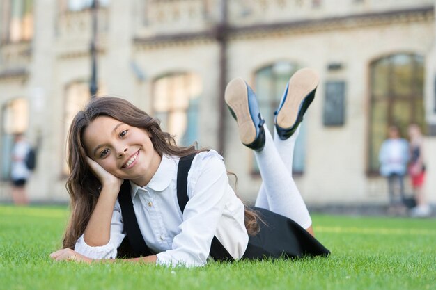 Relax at school yard. Nice time. School break for rest. Happy smiling pupil. Girl cute kid laying green grass. Happy kid relaxing outdoors. Girl school uniform enjoy relax. Cheerful schoolgirl.