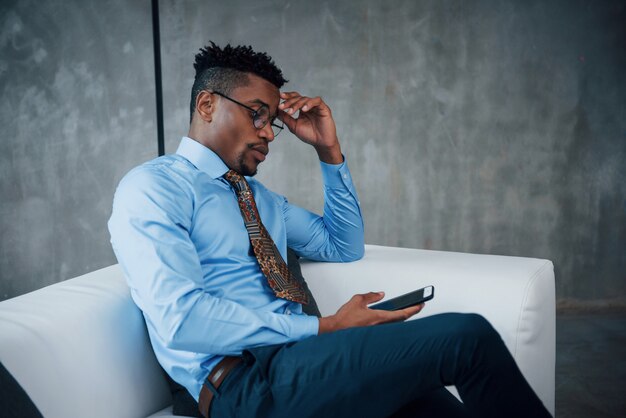 Relax time. Close up portrait of young afro american guy in glasses and classic wear sitting on sofa