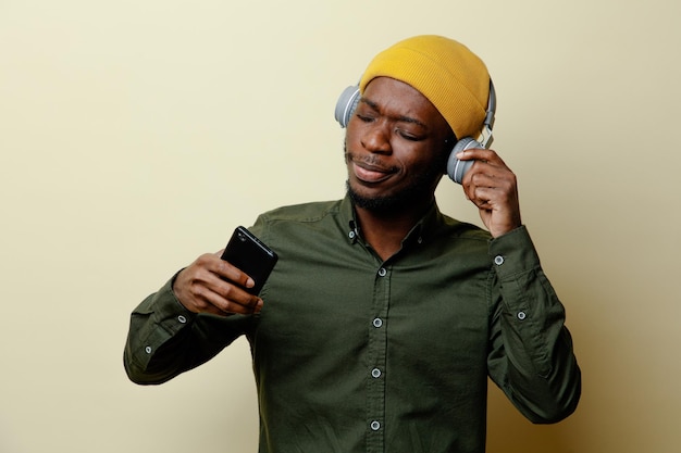 Rilassatevi il giovane maschio afroamericano con cappello e cuffie con in mano il telefono che indossa una camicia verde isolata su sfondo bianco