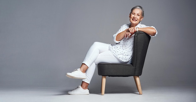 アーンド・イットをリラックスしてください。灰色の背景に椅子に座っている年配の女性のスタジオショット