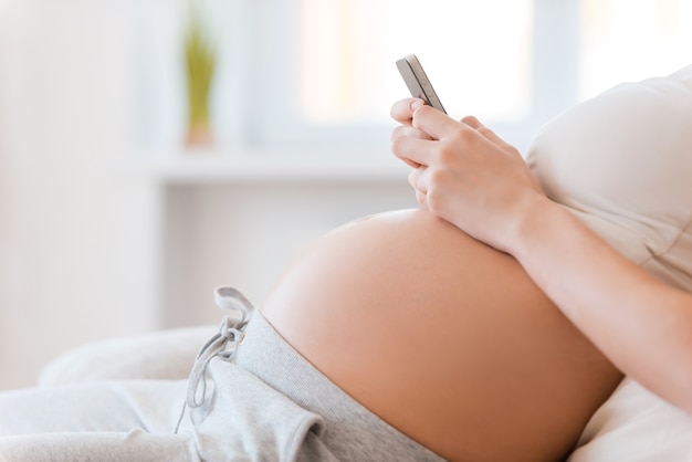 Отдых дома. Крупный план беременной молодой женщины, держащей мобильный телефон, сидя на диване