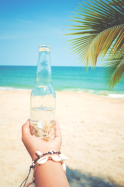 Photo relaxation and leisure in summer - young tanned woman hand holding bottle with seashell at tropical beach in summer. vintage color tone effect