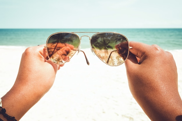 여름의 휴식과 여가 - 여름에 열대 해변에서 선글라스를 든 젊은 그을린 여성의 손. 빈티지한 색조