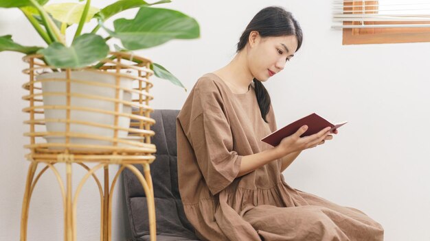 リラクゼーションライフスタイルのコンセプトリビングルームでリラックスするために座って本を読んで若いアジアの女性