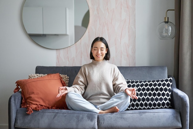 Relaxation and patience smiling young asian woman in cozy room sitting on sofa and meditating doing 