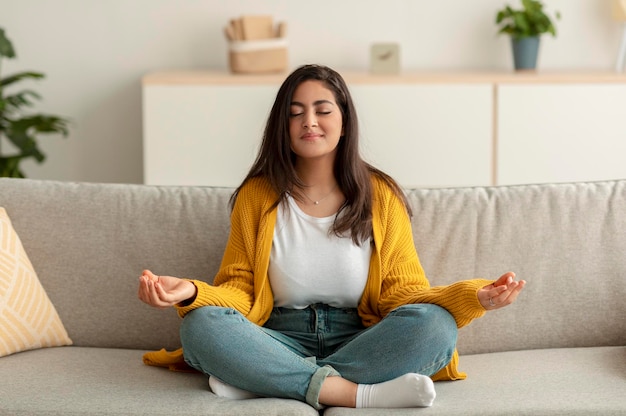 Relaxation stress relief concept Peaceful arab woman sitting in lotus position on couch meditating with closed eyes