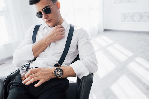 Relaxation with alcohol. Young stylish businessman in sunglasses sitting on a black chair with glass of whiskey.