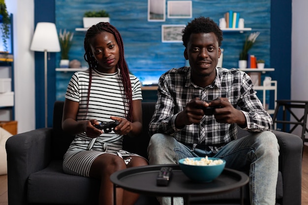 Relaxed african american couple playing video games on console while having a good time sitting at home on the sofa. Joyful people enjoying free time while having fun together in living room.