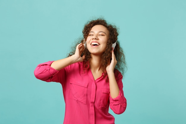 Relaxed african girl in casual clothes keeping eyes closed, listening music with headphones isolated on blue turquoise wall background. People sincere emotions, lifestyle concept. Mock up copy space.