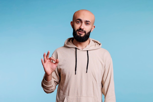 Relaxed arab man showing approval with ok symbol while looking at camera. Calm young bearded person making okay acceptance gesture with fingers while posing for studio portrait