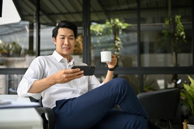 Relaxed Asian businessman watching videos on his phone while enjoying his coffee