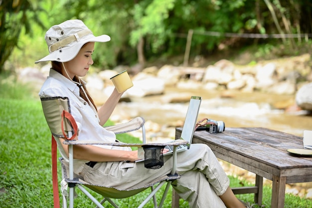川の近くに座っている間コーヒーをすすりながらラップトップコンピューターを使用してリラックスしたアジアの女性キャンピングカー