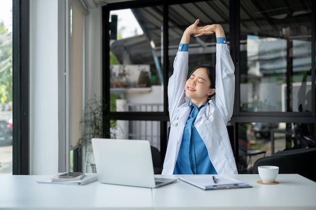 A relaxed Asian female doctor is stretching her arms and taking a break after work at her desk