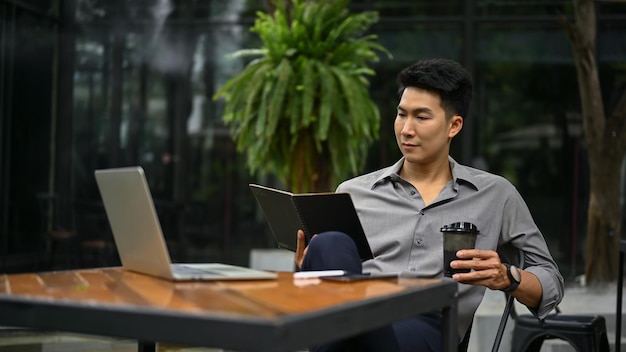 Relaxed Asian man reading a book and sipping coffee at a coffee shop outdoor space