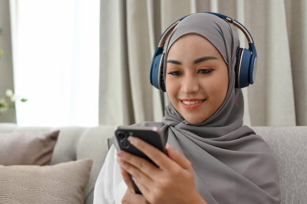 Расслабленная азиатская мусульманка слушает музыку в наушниках и пользуется смартфоном