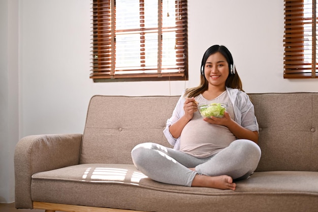 Relaxed Asian pregnant woman eating a healthy salad bowl on sofa in the living room