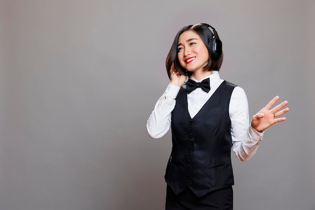 Relaxed asian waitress listening to music in headphones. Smiling cheerful restaurant woman employee wearing uniform enjoying song in wireless earphones while posing in studio