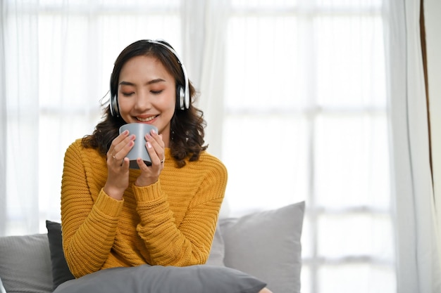 リビングルームで熱いコーヒーやお茶をすすりながら音楽を聴いているリラックスしたアジア人女性