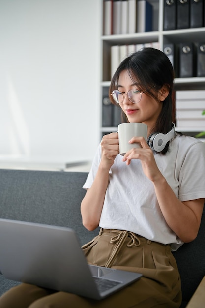 Relaxed Asian woman sits on a sofa in living room smelling her coffee while working from home