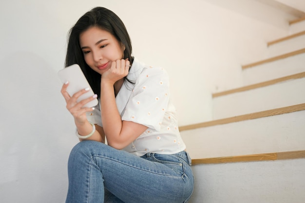 Расслабленная азиатка сидит на лестнице и наслаждается прокруткой своего смартфона в социальных сетях