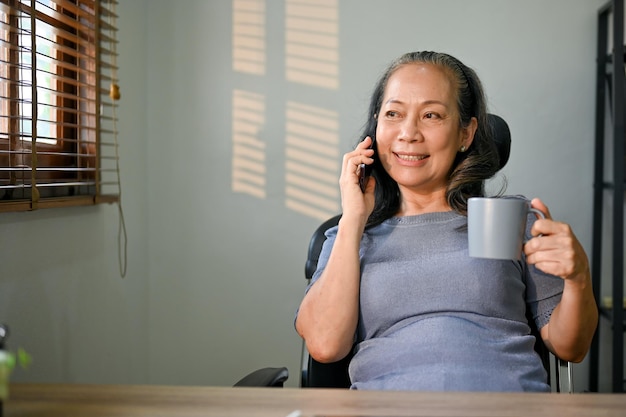 Rilassata imprenditrice asiatica parlando al telefono e sorseggiando caffè alla sua scrivania