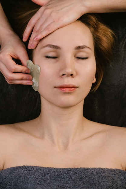 Relaxed beautiful woman receiving facial massage with gua sha stone