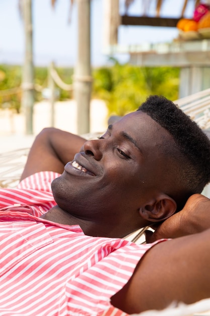 Relaxed biracial man lying in hammock smiling at sunny beach bar. Relaxation, lifestyle, free time, summer and vacation, unaltered.