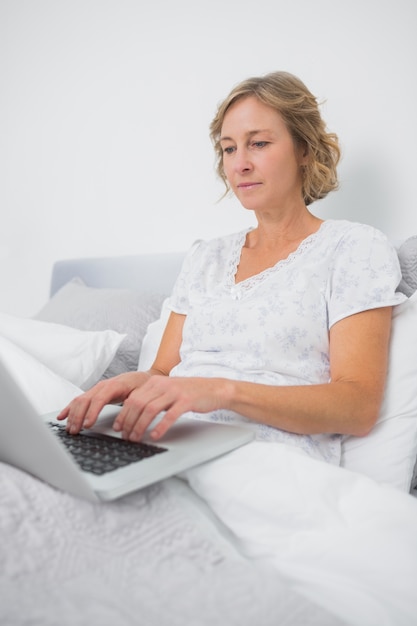 Relaxed blonde woman sitting in bed using laptop 