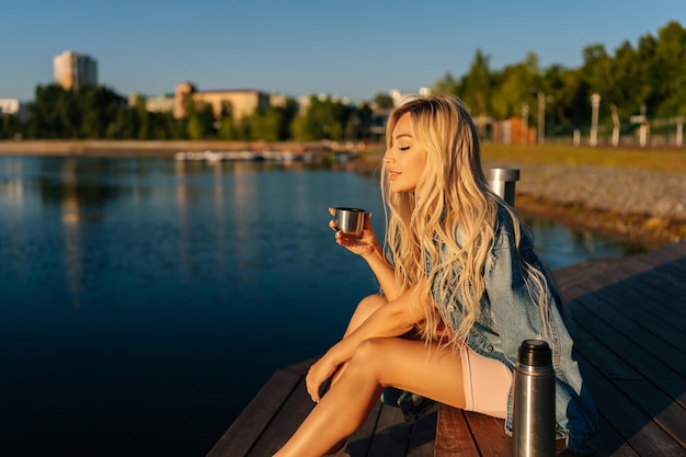 Relaxed blonde young woman holding cup with hot coffee from thermos sitting on beach by wooden pier on summer sunny morning