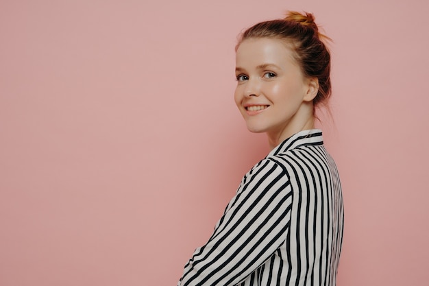 Relaxed brunette teenage girl dressed in white and black shirt with hair in bun, looking at camera with crossed arms while smiling, feeling happy while standing alone in front of pink background