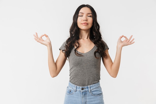 relaxed brunette woman 20s dressed in basic clothes meditating and gesturing with fingers isolated over white wall